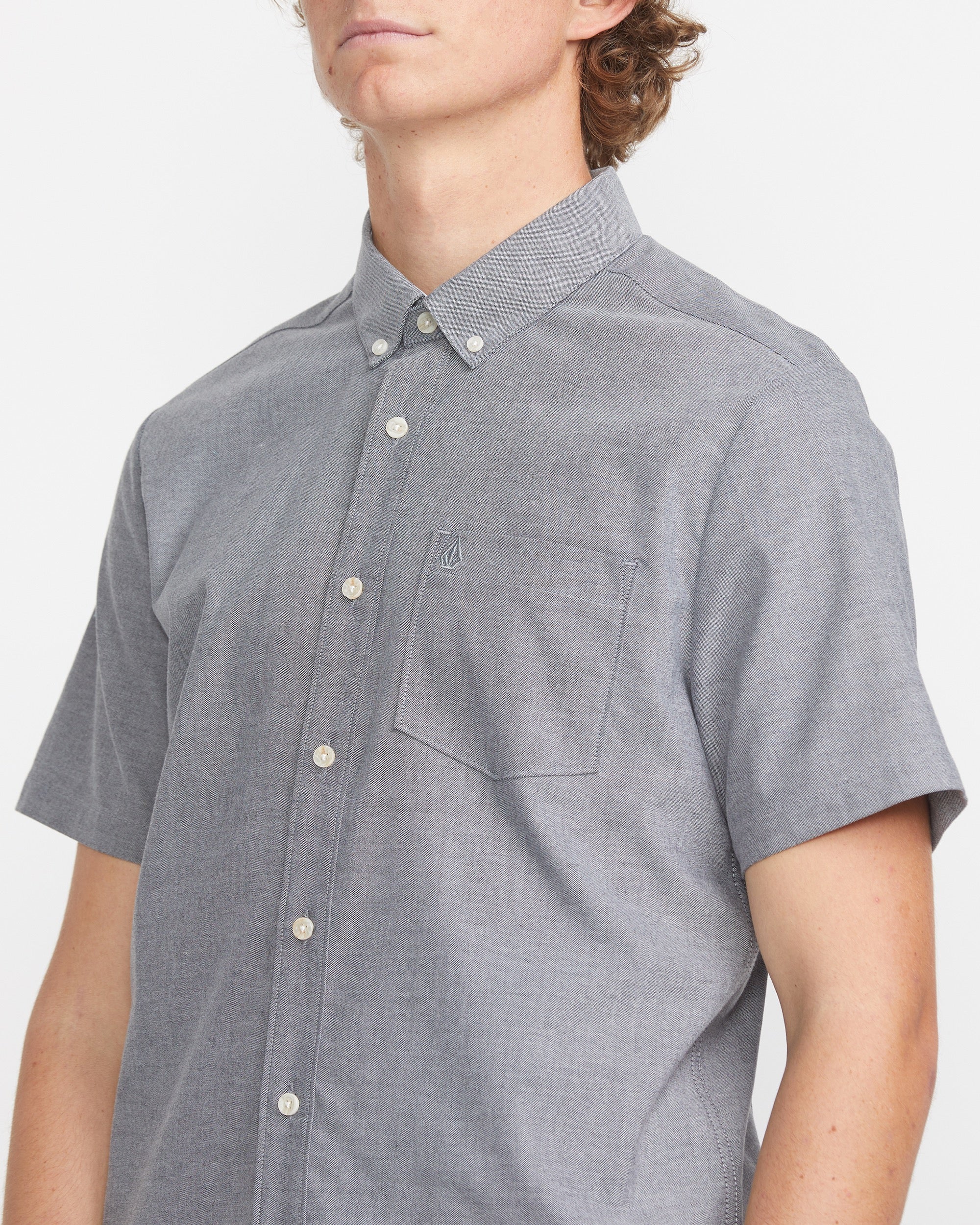 A person in the Volcom Men's Everett Oxford Short Sleeve Tee, featuring a chest pocket and subtle branding, stands against a white background. The slim fit and modern design complement their slightly turned, partially visible head.
