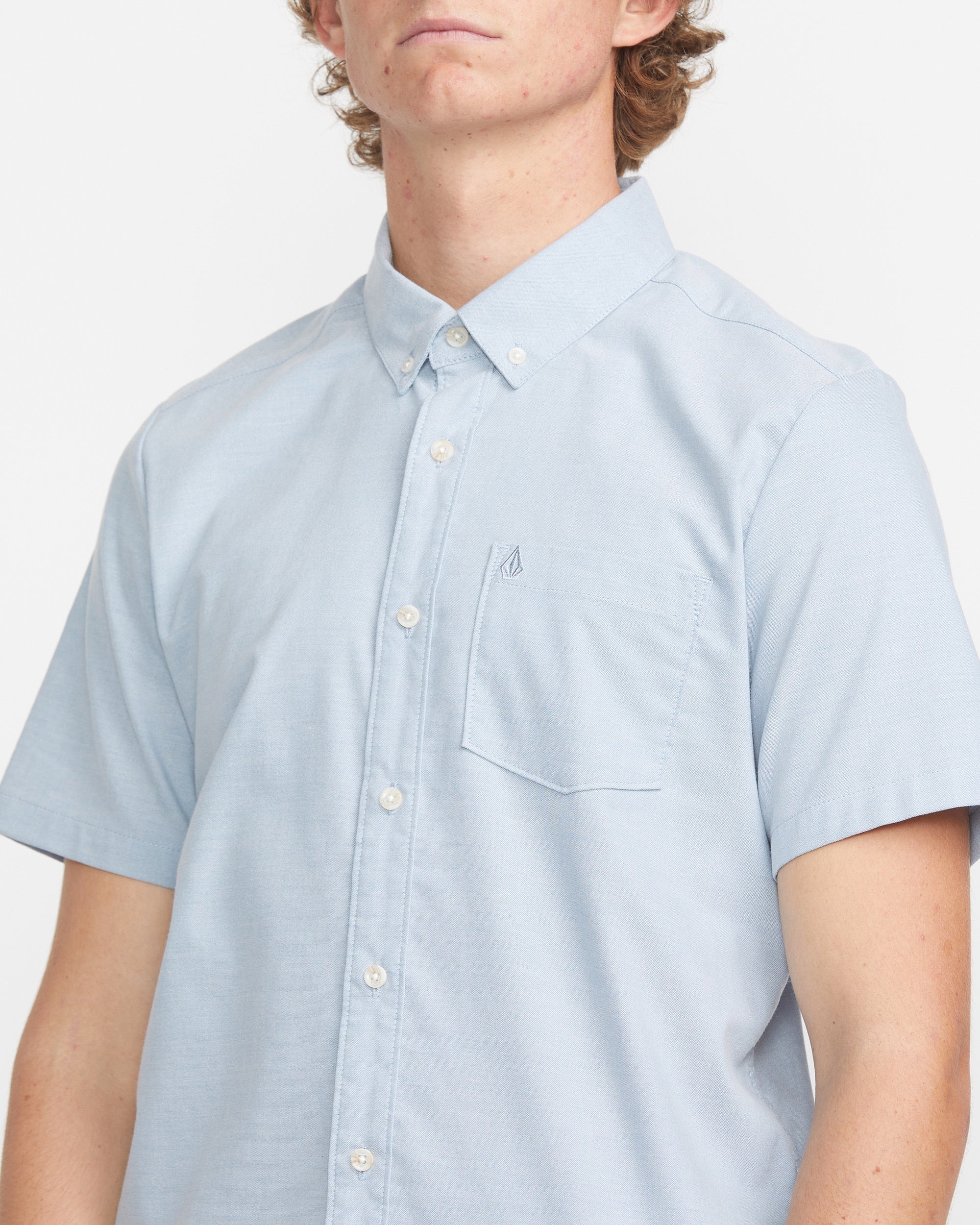 A person is seen wearing the Volcom Men's Everett Oxford Short Sleeve shirt in light blue, featuring a slim fit and breast pocket, standing against a plain white background with their face partially out of frame.