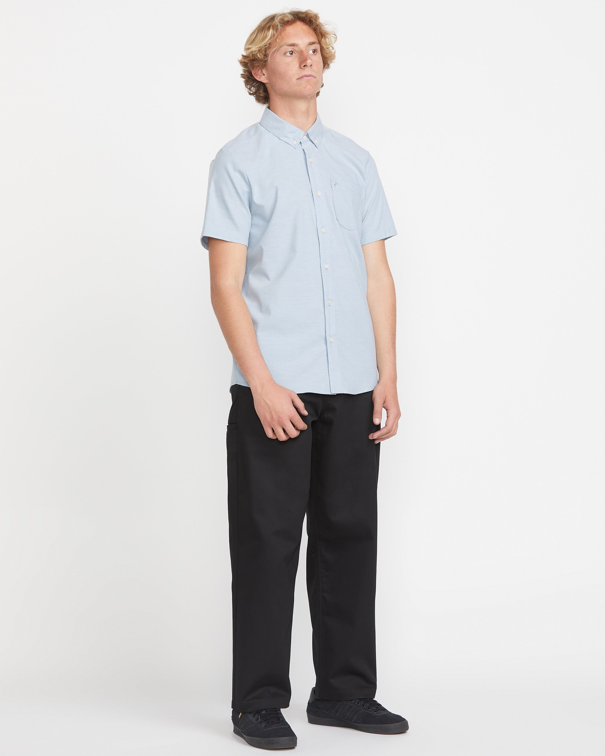 A young man with blond hair stands against a white background, wearing the Volcom Men's Everett Oxford Short Sleeve shirt in light blue, paired with black pants and shoes, embodying a modern mix of classic and contemporary style.