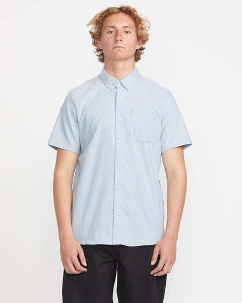 A young man with curly blond hair wears the Volcom Men's Everett Oxford Short Sleeve in light blue and black pants, standing against a plain white background, looking straight at the camera.