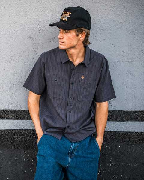 A young man, wearing the Volcom Men’s Kap Out Workwear Short Sleeve Shirt—featuring a dark, durable design with chest pockets—and blue jeans, leans against a gray wall. He completes the look with a black cap, gazing to his left with his hands in his pockets.