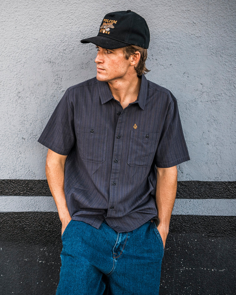 A young man, wearing the Volcom Men’s Kap Out Workwear Short Sleeve Shirt—featuring a dark, durable design with chest pockets—and blue jeans, leans against a gray wall. He completes the look with a black cap, gazing to his left with his hands in his pockets.