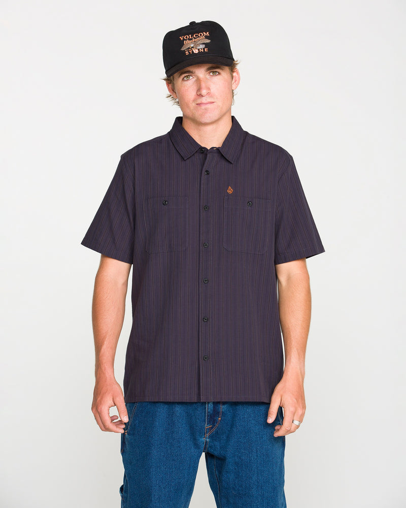 A person wearing the Volcom Men's Kap Out Workwear Short Sleeve Shirt in black, paired with blue jeans and a black cap featuring orange text, stands against a plain white background facing the camera with a neutral expression.