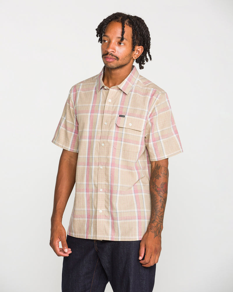 A man with short dreadlocks wears the Volcom Men's Ledges Short Sleeve Shirt, a yarn-dyed plaid classic-fit button-up featuring a front pocket, while standing against a plain white background.