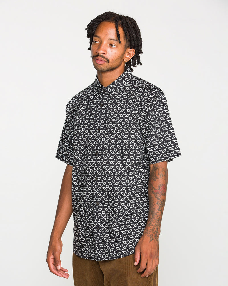 A man with short dreadlocks and tattoos on his arm wears the Volcom Men's Scaler Stone Short Sleeve Shirt and brown pants, standing against a plain white background.