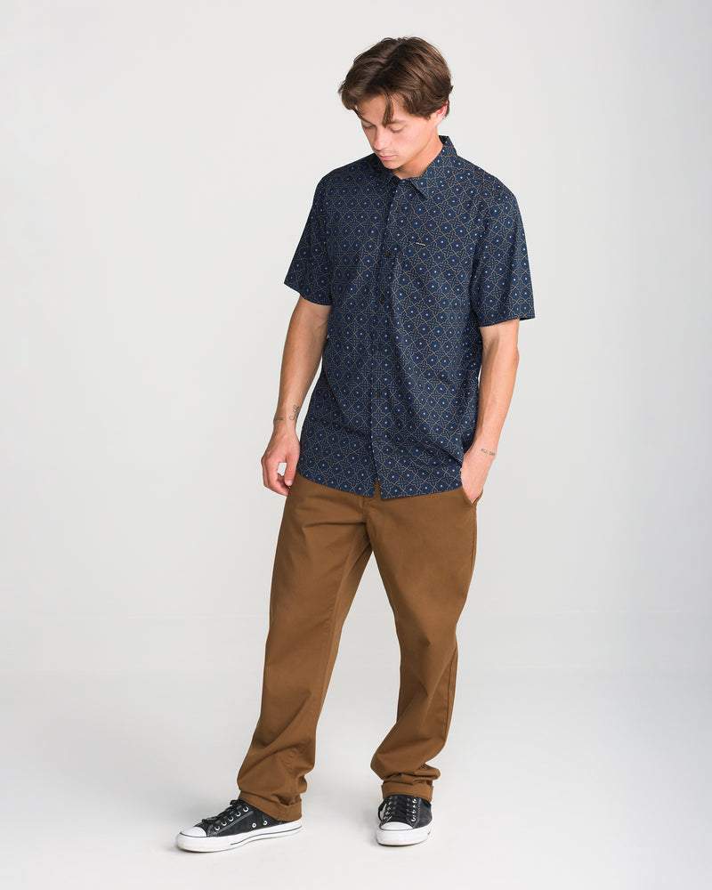 A person wearing a navy blue, geometric-patterned Men’s Scaler Stone Short Sleeve button-up shirt with brown trousers and black sneakers.