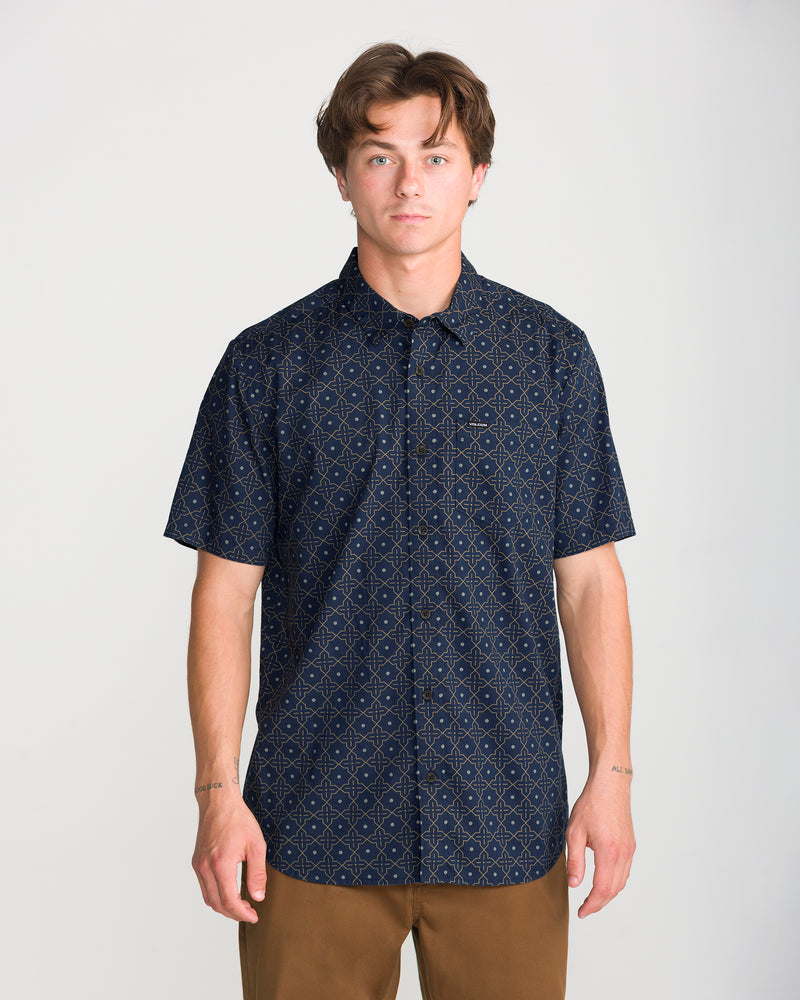 A man wearing a Men’s Scaler Stone Short Sleeve button-up shirt with an all-over dark blue geometric pattern and brown pants.