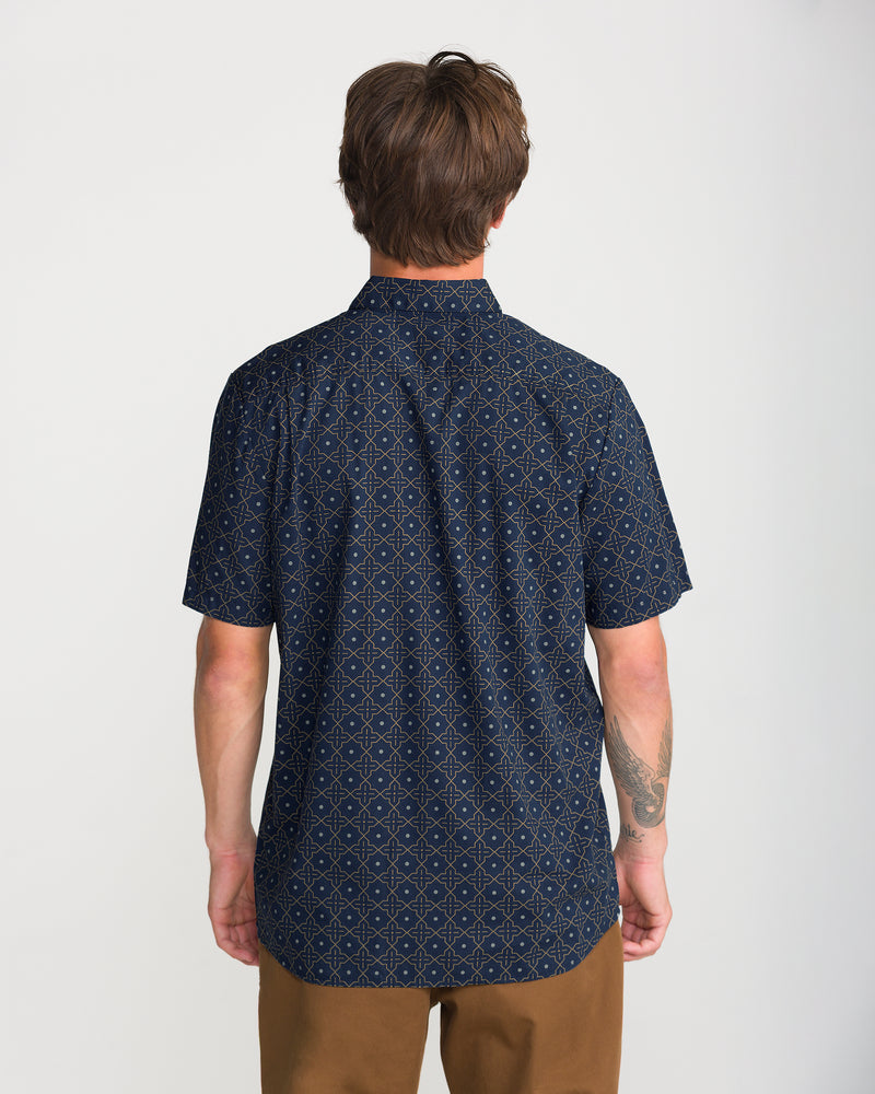 A person seen from the back wearing a Men’s Scaler Stone Short Sleeve, a dark navy Volcom short-sleeve button-up with a repeating tan geometric all-over print.