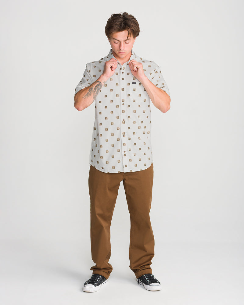 A person wearing a Volcom Men’s Scaler Stone Short Sleeve in off-white with a small geometric print, paired with brown pants and black sneakers.