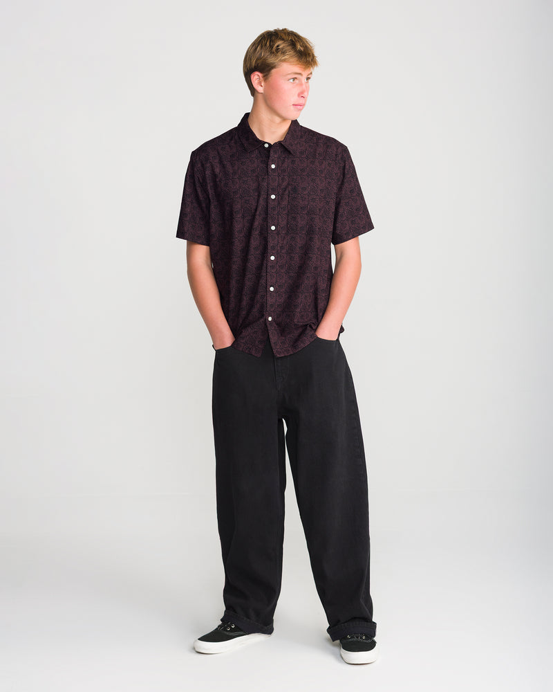 A person stands with hands in pockets wearing a Volcom Men’s Branton Short Sleeve in a boxy-fit, short-sleeve button-down style with an allover print, paired with black loose-fit pants and black sneakers.