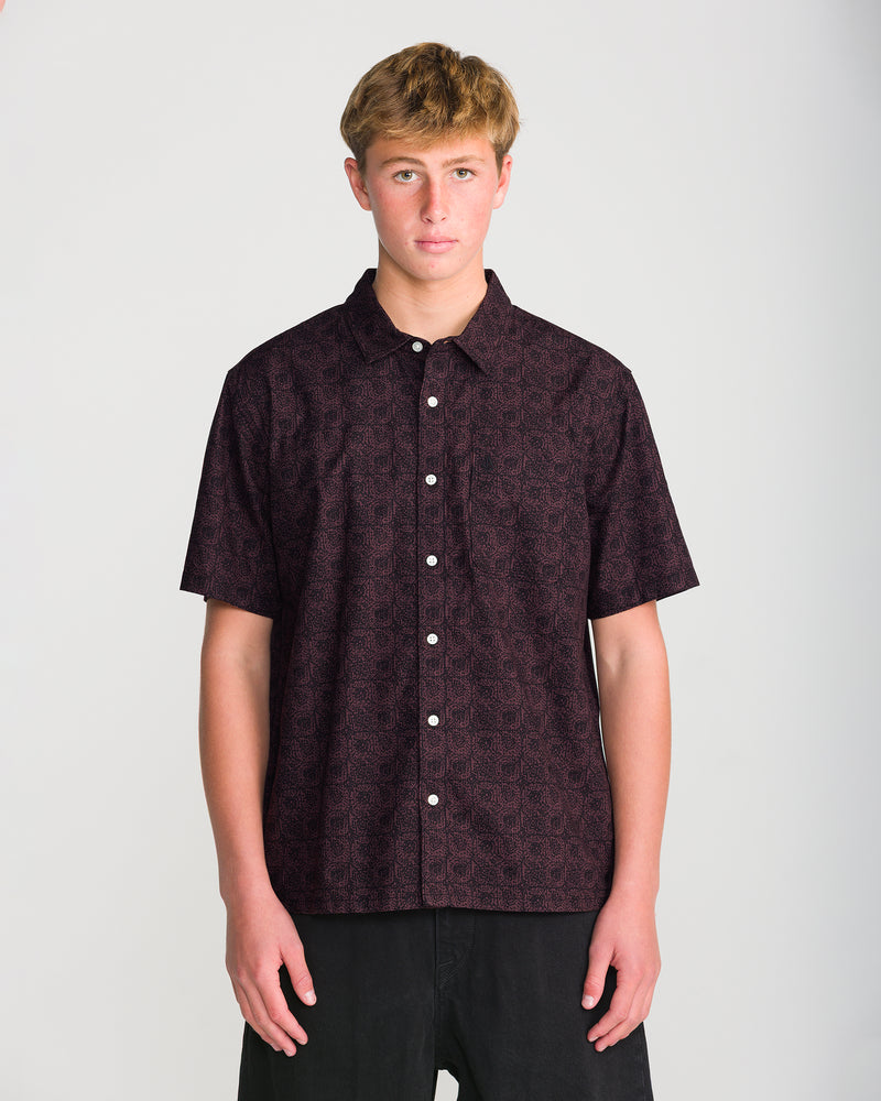 A man wears a Volcom Men’s Branton Short Sleeve, a boxy-fit, short-sleeve button-up shirt with an allover geometric print, paired with black pants.