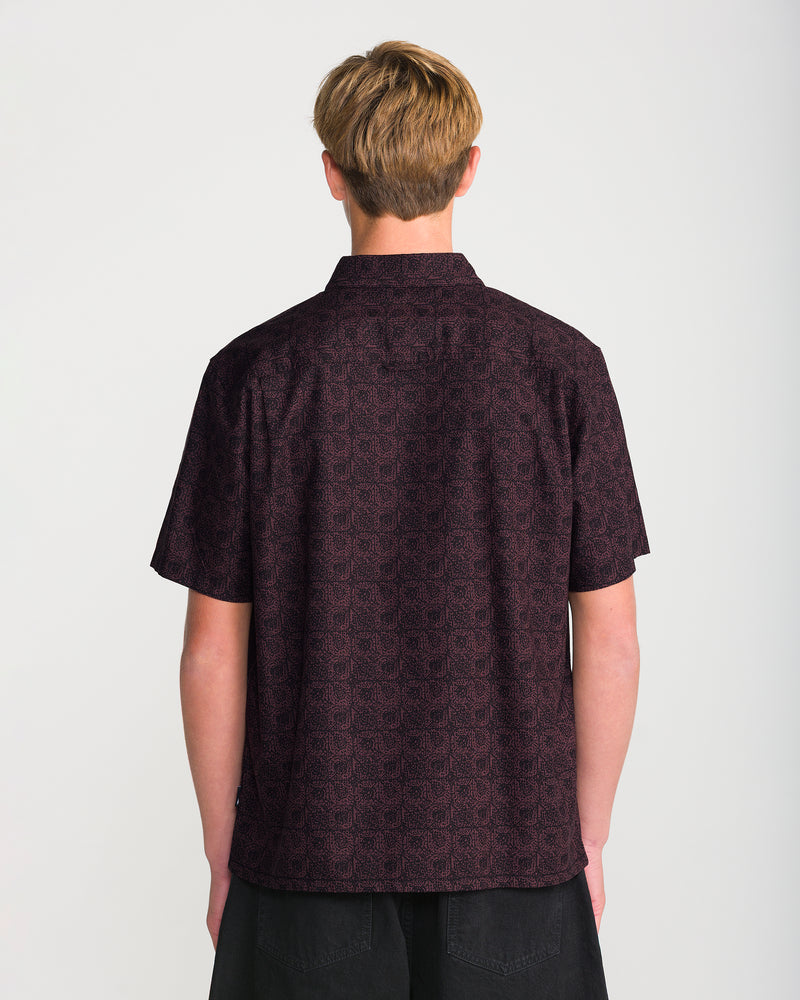 A man seen from behind wearing the Volcom Men’s Branton Short Sleeve, a boxy fit, short-sleeve collared shirt with an allover dark, repeating geometric pattern.
