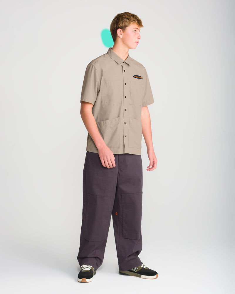 A young man wearing a tan Volcom Men’s Driggs Workwear Short Sleeve button-up shirt with two chest pockets, dark gray loose-fit pants, and sneakers.