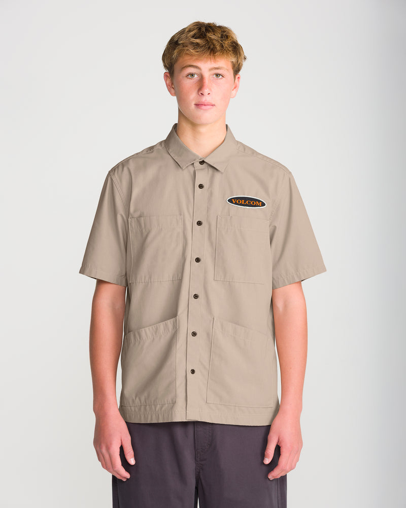 A man wearing a Volcom Men’s Driggs Workwear Short Sleeve, a sturdy plain-weave, tan short-sleeve button-down shirt with a black chest pocket patch in a classic fit.