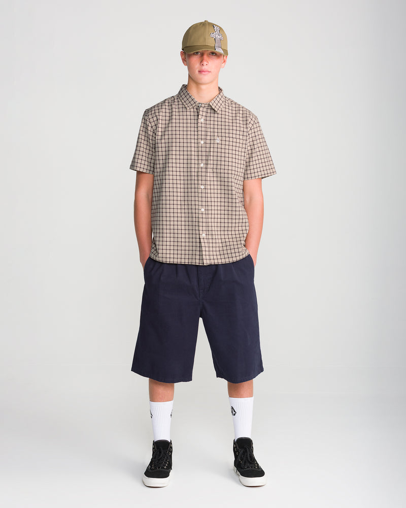 A person wearing a tan plaid Volcom Men’s Flatlands Short Sleeve shirt with a modern boxy fit, navy blue shorts, white socks, black sneakers, and a tan baseball cap.