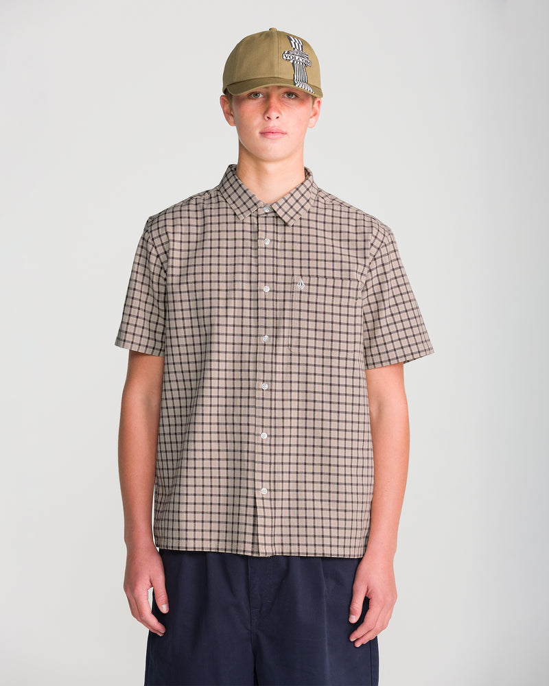 A man wears a Volcom Men’s Flatlands Short Sleeve in a modern boxy fit tan plaid, paired with navy shorts and an olive green baseball cap.