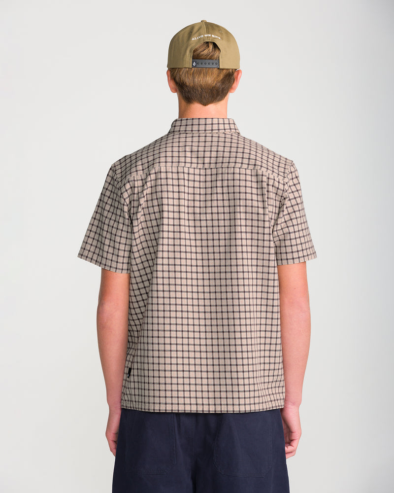 Back view of a person wearing a Volcom Men’s Flatlands Short Sleeve, a modern, boxy-fit beige short-sleeve button-down shirt with a dark brown grid pattern, paired with navy shorts and a tan baseball cap.