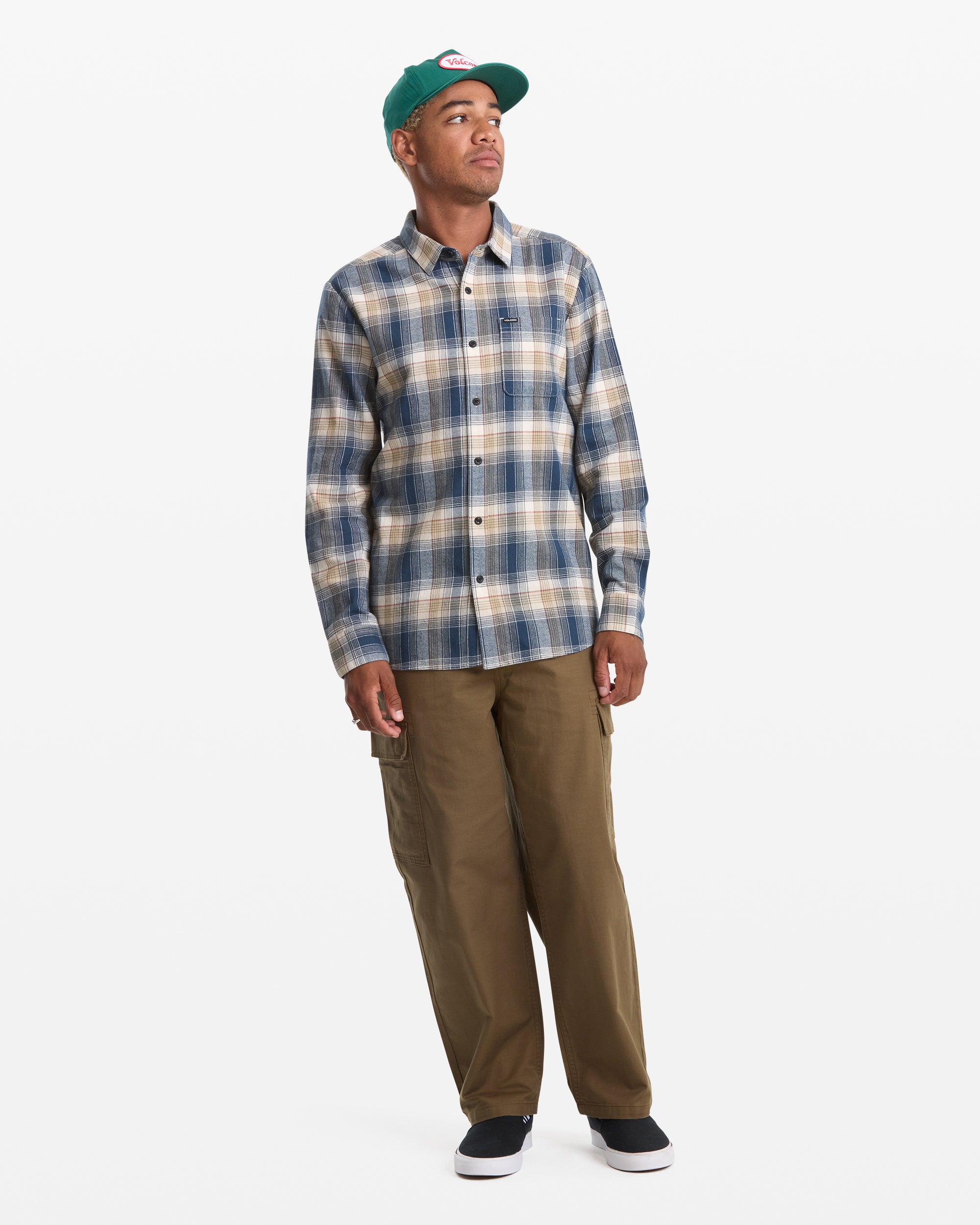 A person wearing a green cap, Volcom Men's Caden Plaid Long Sleeve shirt in blue and beige, brown pants, and black slip-on shoes stands against a plain white background.