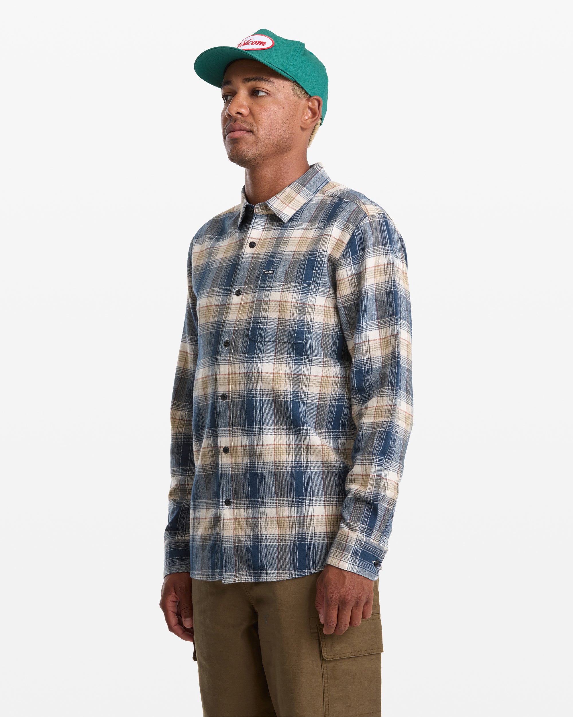 A man stands against a white background wearing a green cap, brown pants, and the Volcom Men's Caden Plaid Long Sleeve shirt. He is facing slightly to the left with a neutral expression.
