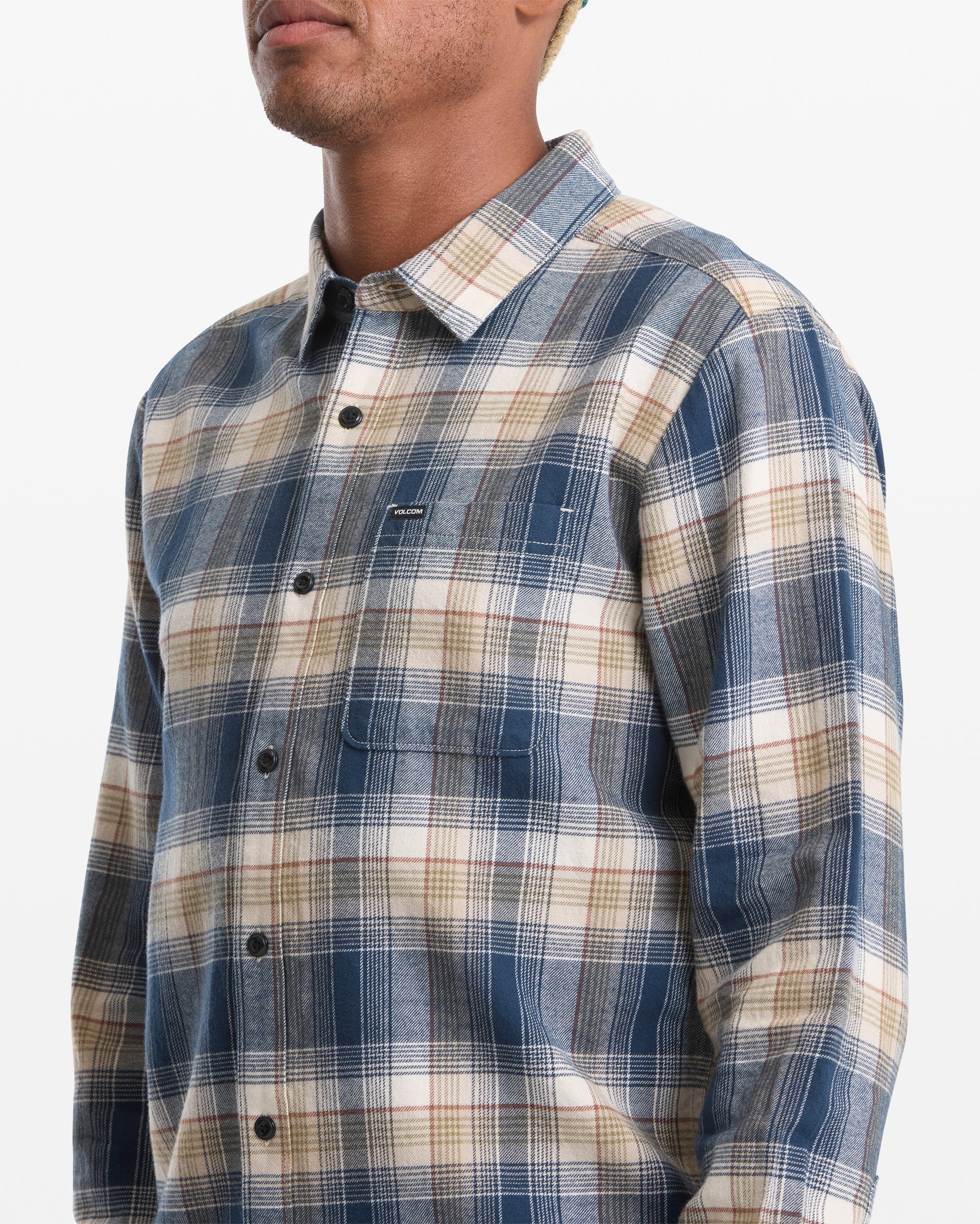A man wears the Volcom Men's Caden Plaid Long Sleeve shirt—blue, beige, and white cotton flannel with a front pocket—shown from the chest up against a plain white background.