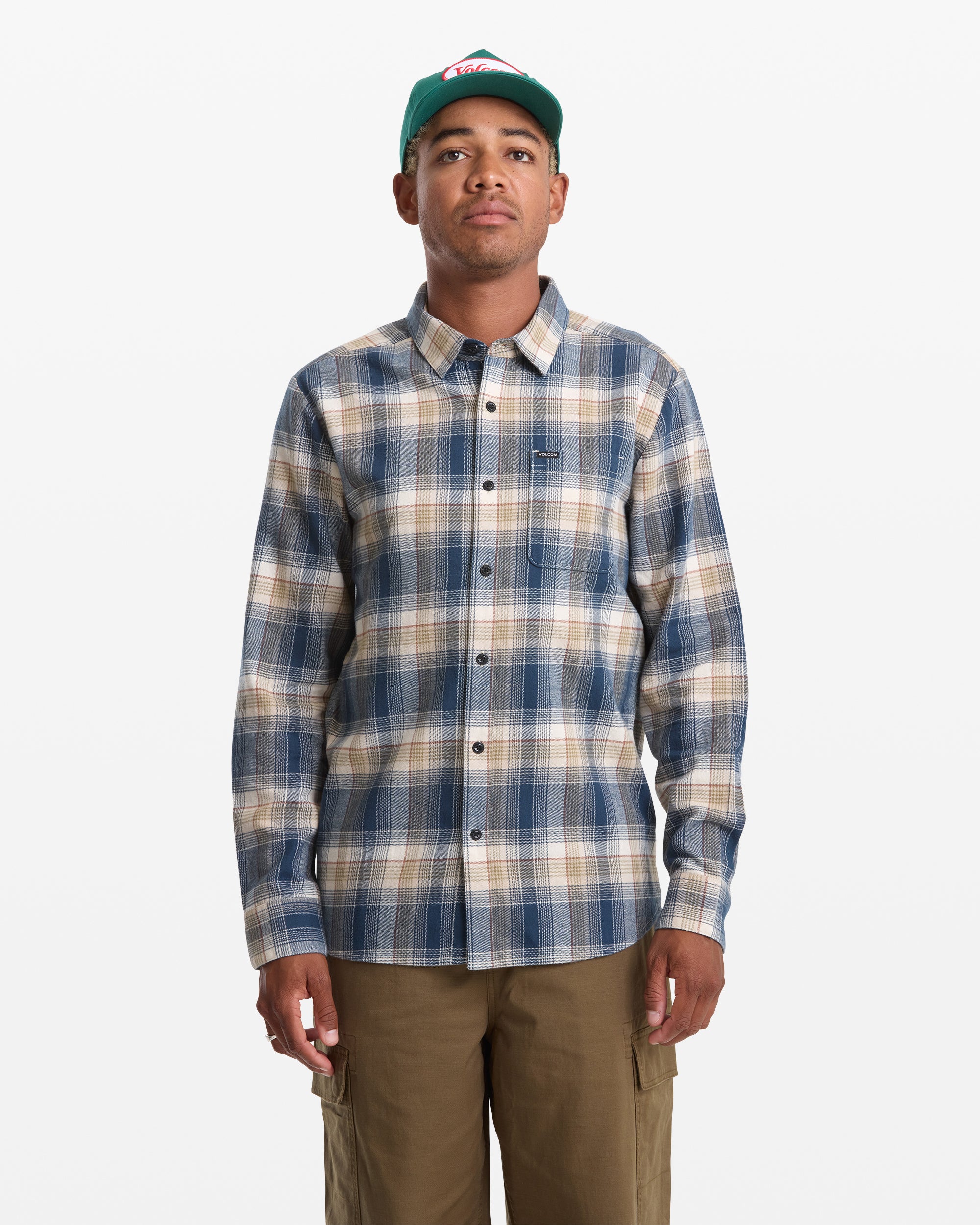 A person wearing a green baseball cap, khaki cargo pants, and the Volcom Men's Caden Plaid Long Sleeve shirt in blue and beige stands against a white background.