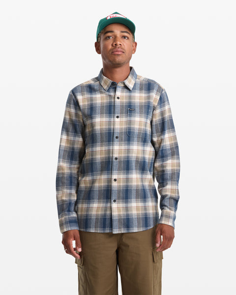 A person wearing a green baseball cap, khaki cargo pants, and the Volcom Men's Caden Plaid Long Sleeve shirt in blue and beige stands against a white background.