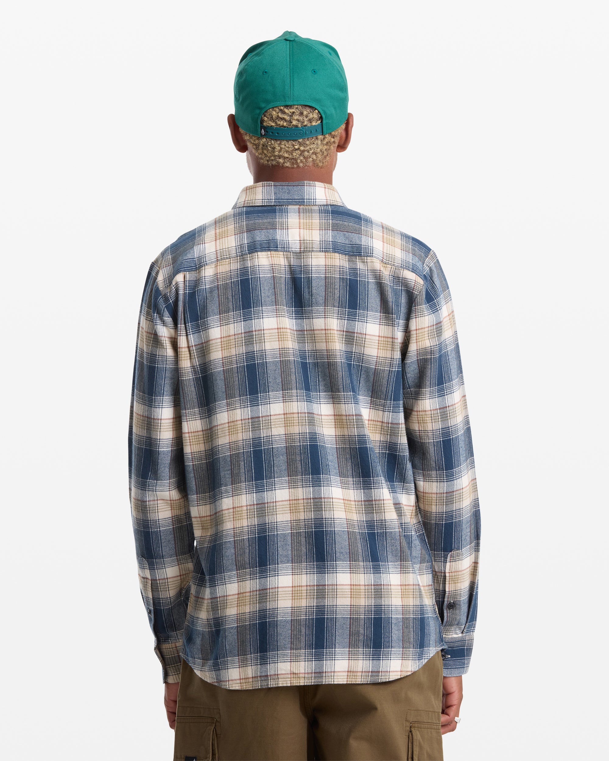 A person wearing a green cap, khaki pants, and a Volcom Men's Caden Plaid Long Sleeve shirt in blue and beige stands with their back facing the viewer.