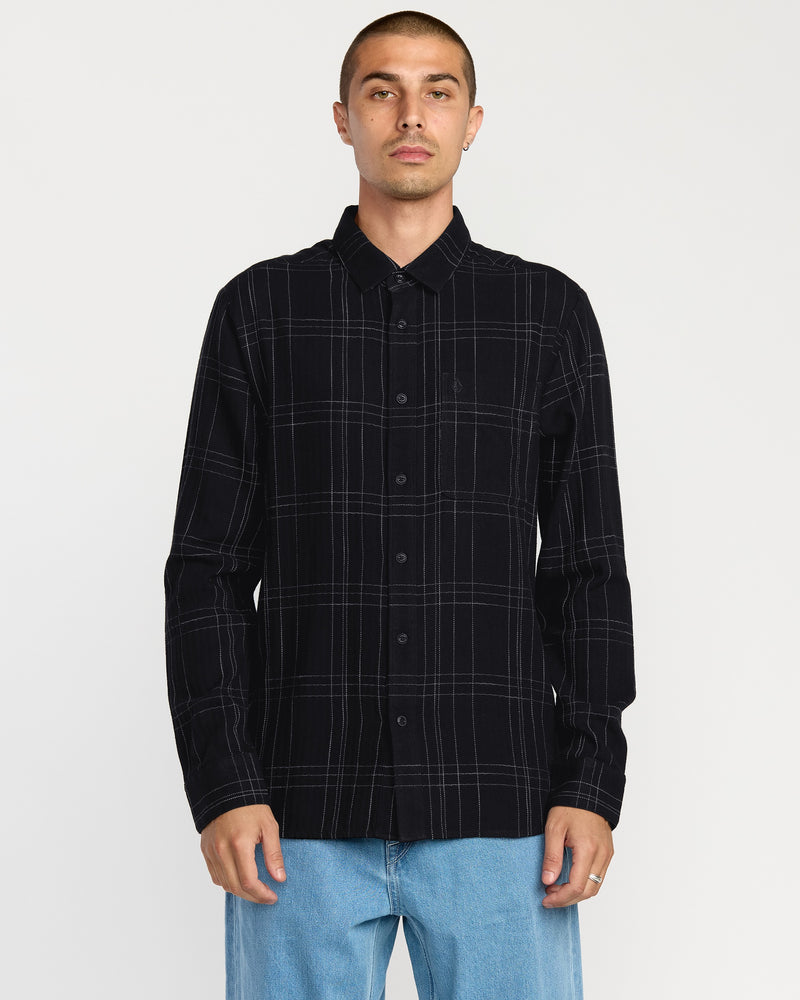 A man stands facing the camera against a plain white background, wearing a Volcom Men's Date Knight Long Sleeve Shirt in black and white plaid, paired with light blue jeans.