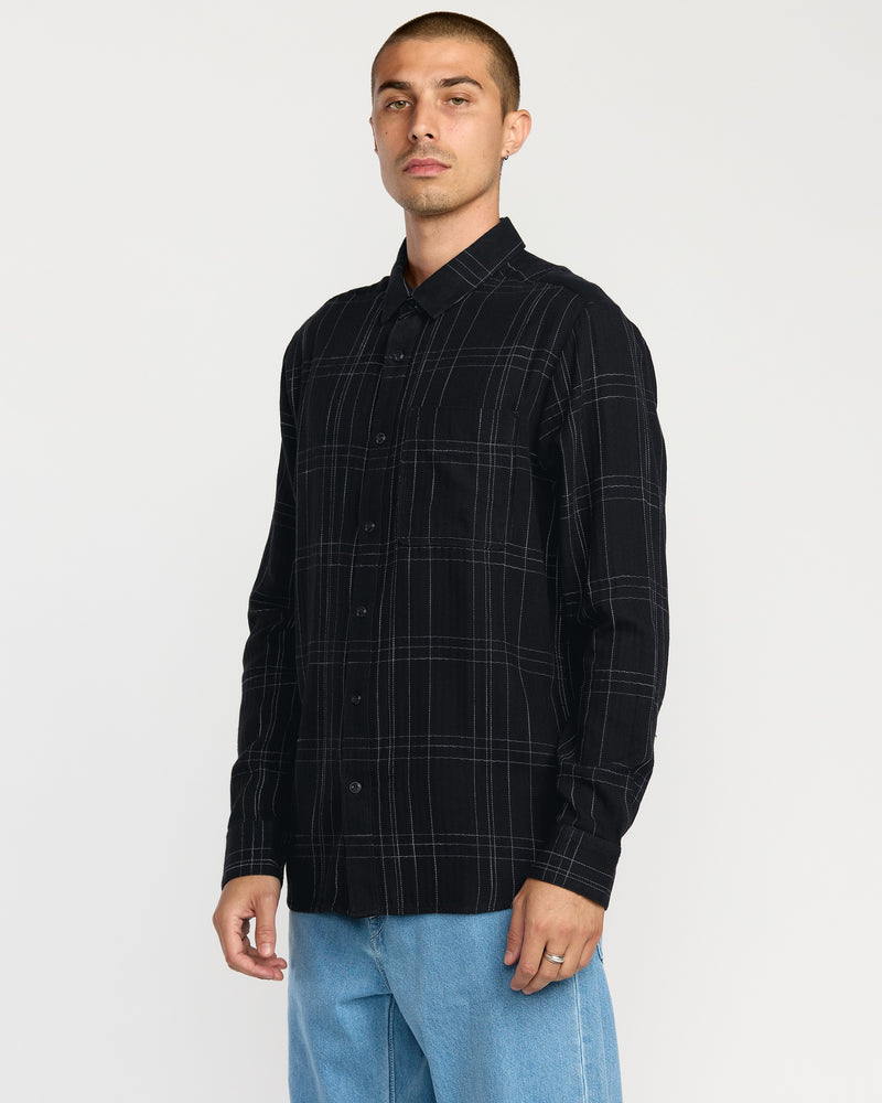 A man wearing Volcom's Men's Date Knight Long Sleeve Shirt, a classic-fit black shirt with white plaid lines made from a premium cotton blend, pairs it with light blue jeans while standing against a plain white background.