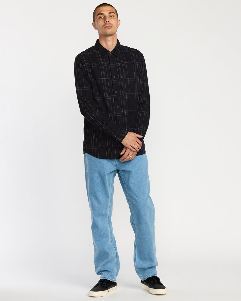 A person stands against a plain white background, wearing the Volcom Men's Date Knight Long Sleeve Shirt and light blue jeans, paired with black sneakers with white soles. Their arms are relaxed, with one hand clasping the other wrist.