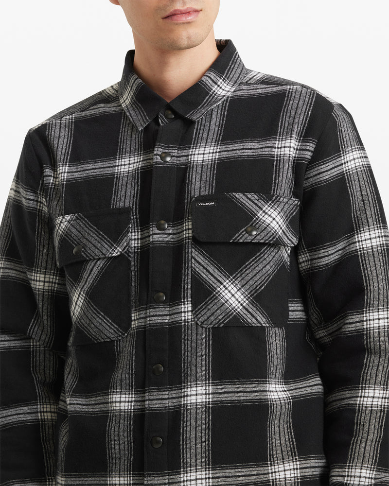 A man is shown from shoulders to waist against a plain white background, wearing the Volcom Men's Wilderstone Long Sleeve shirt—a black and white plaid cotton woven top with two chest pockets.