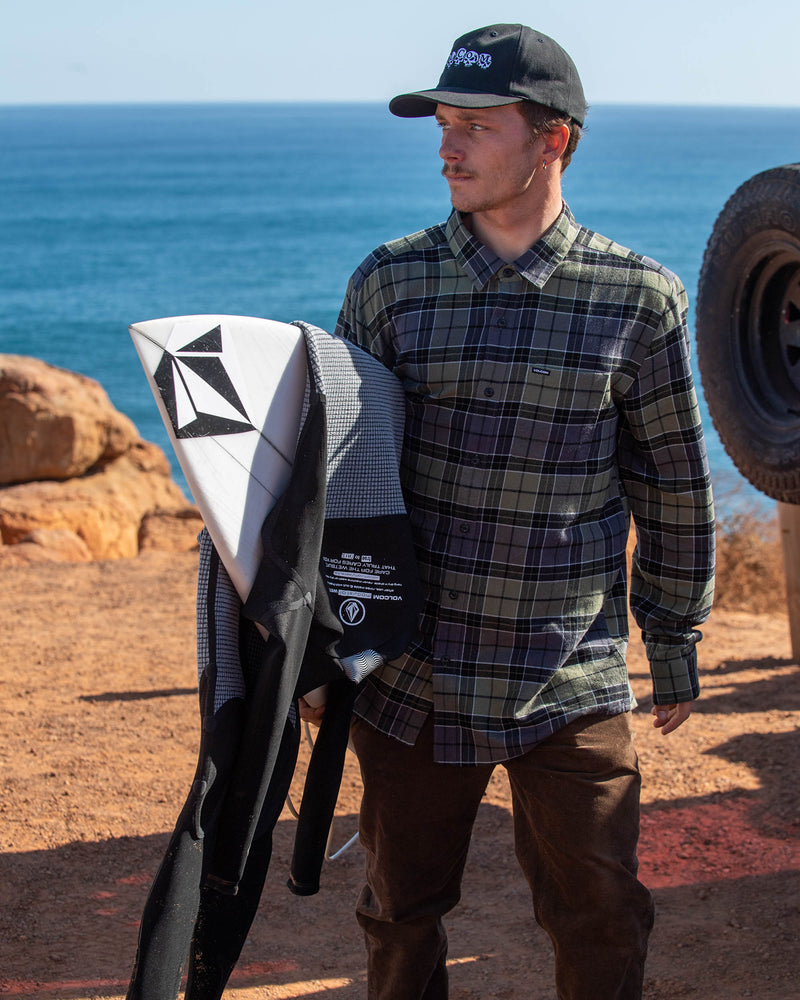 A man wearing the Volcom Men's Levelstone Long Sleeve Shirt, brown pants, and a black cap stands on a rocky coastal path holding a surfboard and wetsuit, with the ocean in the background.