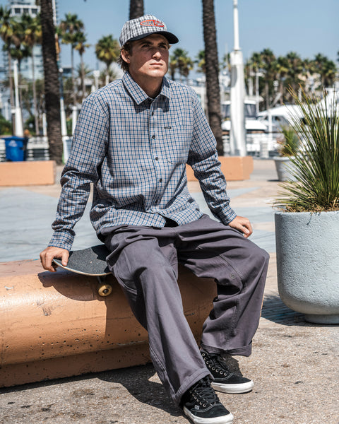 Wearing the Volcom Men’s Landos Long Sleeve Shirt, which features a yarn-dyed plaid pattern, oversized chest pocket, and relaxed boxy silhouette, a person sits on a concrete barrier holding a skateboard. Palm trees and a marina are visible in the sunny background.
