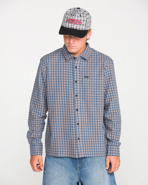 A person wearing the Volcom Men's Landos Long Sleeve Shirt, featuring a checked pattern and an oversized chest pocket, stands against a plain white background looking down. They pair the shirt with light-colored jeans, a plaid baseball cap, and have their hands at their sides.
