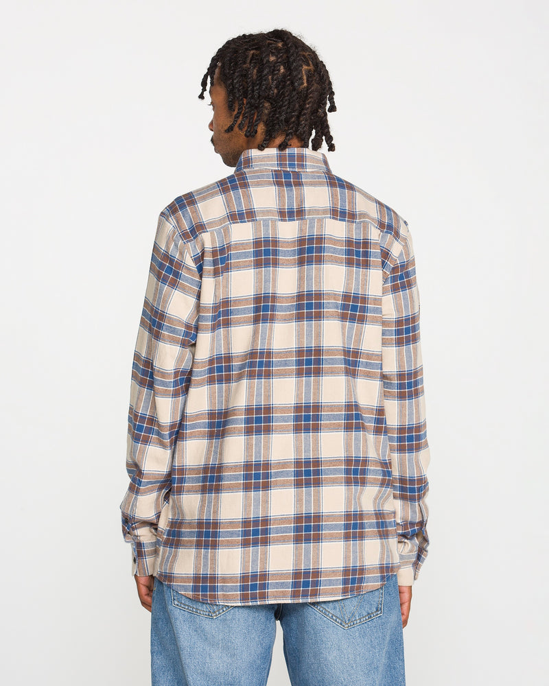 A person with short dreadlocks is shown from the back, wearing the Volcom Men's Caden Plaid Long Sleeve Shirt in beige, blue, and brown, paired with light blue jeans against a plain white background.