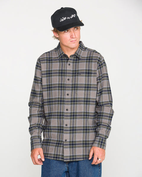 A man models the Volcom Men's Caden Plaid Long Sleeve Shirt in classic fit gray and dark blue, paired with a black cap and blue jeans, standing against a plain white background and looking slightly to the side.