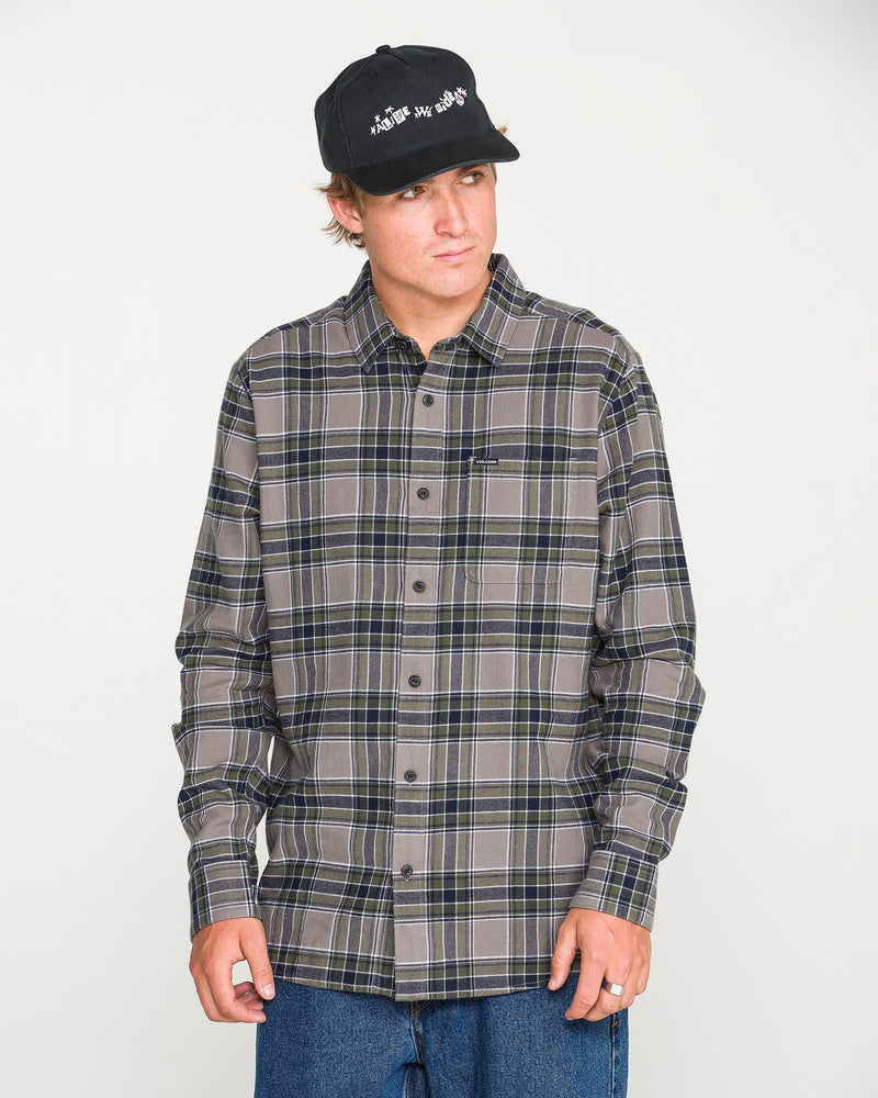 A man models the Volcom Men's Caden Plaid Long Sleeve Shirt in classic fit gray and dark blue, paired with a black cap and blue jeans, standing against a plain white background and looking slightly to the side.