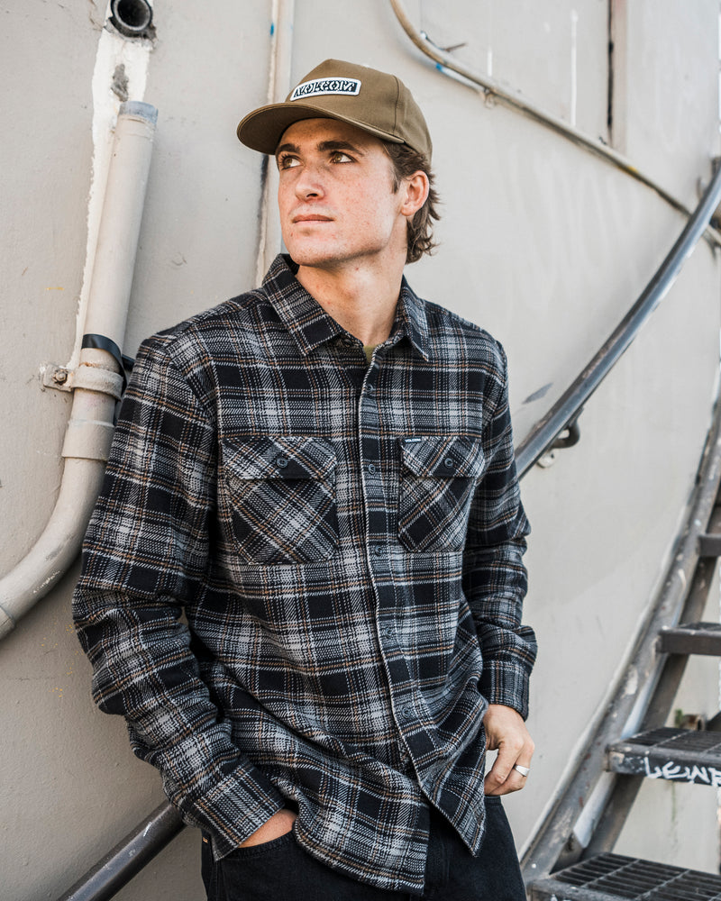 Wearing the Volcom Men’s Johnstone Long Sleeve Shirt, a man with a relaxed, boxy fit and tan cap leans against a wall lined with pipes, gazing to the side with his hands in his pockets and stairs behind him.