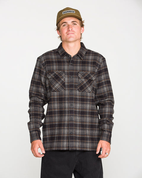 A person wearing a beige cap, black pants, and the Volcom Men's Johnstone Long Sleeve Shirt — a black-and-brown plaid heavyweight flannel with chest pockets — stands facing forward against a plain white background.