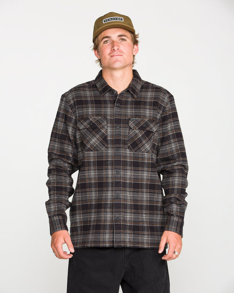 A person wearing a beige cap, black pants, and the Volcom Men's Johnstone Long Sleeve Shirt — a black-and-brown plaid heavyweight flannel with chest pockets — stands facing forward against a plain white background.