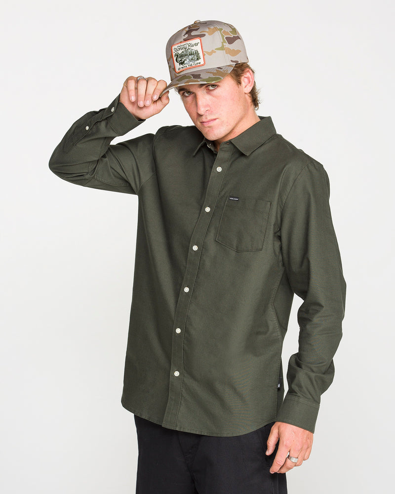 A person wearing the Volcom Men's Veeco Oxford Long Sleeve Shirt in Climbing Ivy green and black pants adjusts a camouflage baseball cap. The classic fit and micro suede fabric of the shirt add a subtle touch of sophistication against the plain white background.
