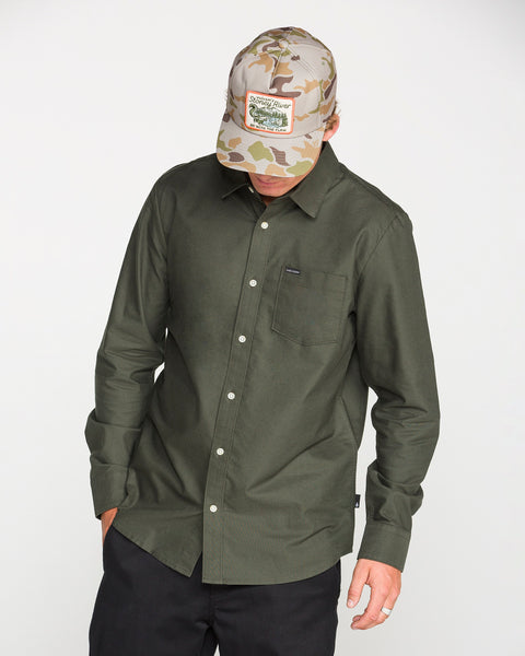 A person wearing the Volcom Men's Veeco Oxford Long Sleeve Shirt in Climbing Ivy green, paired with black pants and a camouflage baseball cap with a label, looks down with one hand in a pocket against a plain light background.