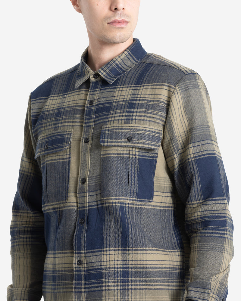 A man wears the Volcom Men's Parkstone Long Sleeve Shirt, a blue and beige plaid button-up with two chest pockets, standing against a plain light background.