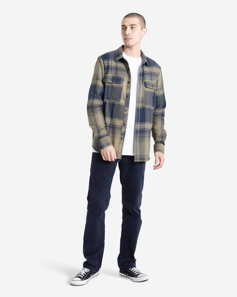 A man stands slightly turned, wearing the Volcom Men's Parkstone Long Sleeve Shirt—a beige and navy plaid button-up—over a white t-shirt, with dark pants and black low-top sneakers, set against a plain white background.