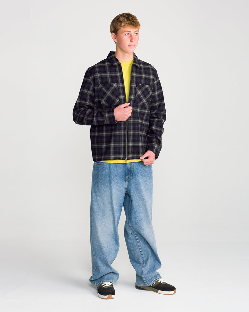 A person wears a Volcom Men’s Fieldstone Long Sleeve in a black and white plaid with a modern boxy fit over a yellow t-shirt, paired with baggy blue jeans and sneakers.