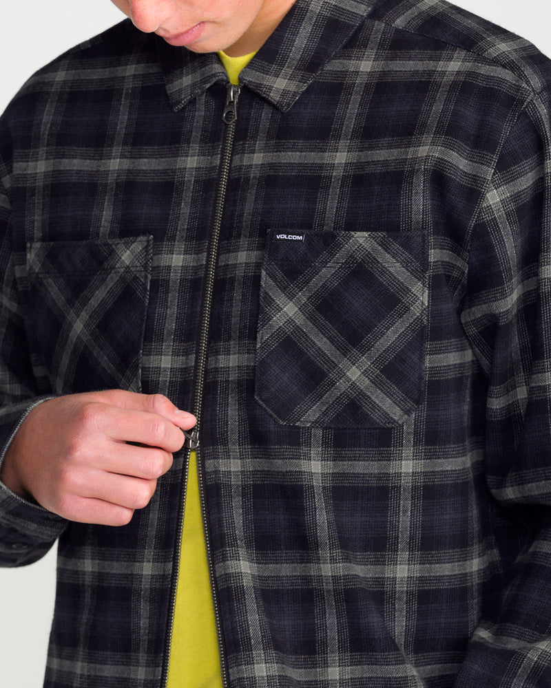 A person wearing a heavyweight, grey and black plaid Men’s Fieldstone Long Sleeve over a yellow shirt, holding the front zipper.