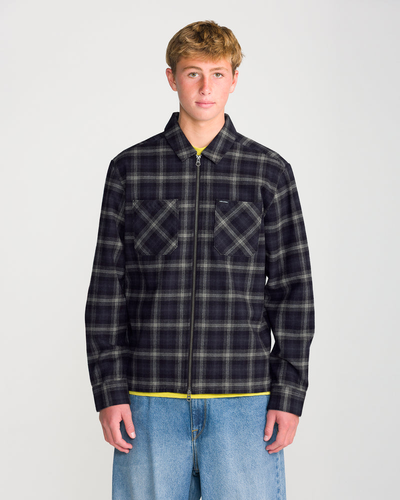 A young man wears a Volcom Men’s Fieldstone Long Sleeve in a black and grey modern boxy fit over a yellow shirt and blue jeans.