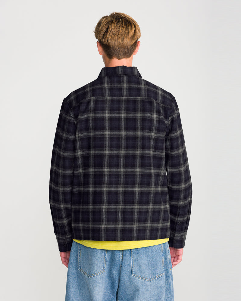 Back view of a person wearing a Volcom Men’s Fieldstone Long Sleeve, a boxy fit black and grey plaid heavyweight flannel shirt over a yellow shirt and blue jeans.