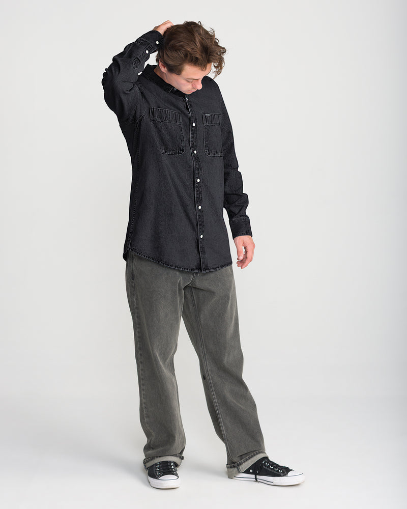 A person wearing a black Men’s Goodwin Long Sleeve shirt with two chest pockets, loose-fitting grey jeans, and black low-top sneakers.