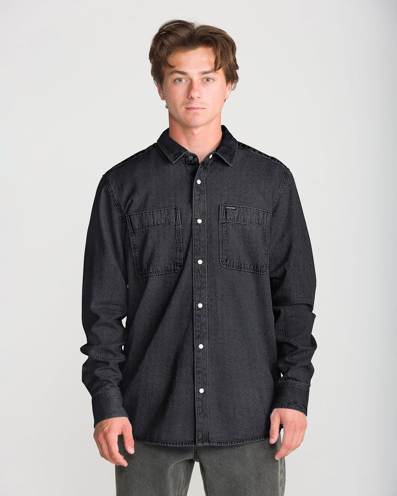A man wearing a black Men’s Goodwin Long Sleeve relaxed fit midweight denim shirt with two chest pockets and dark denim pants.